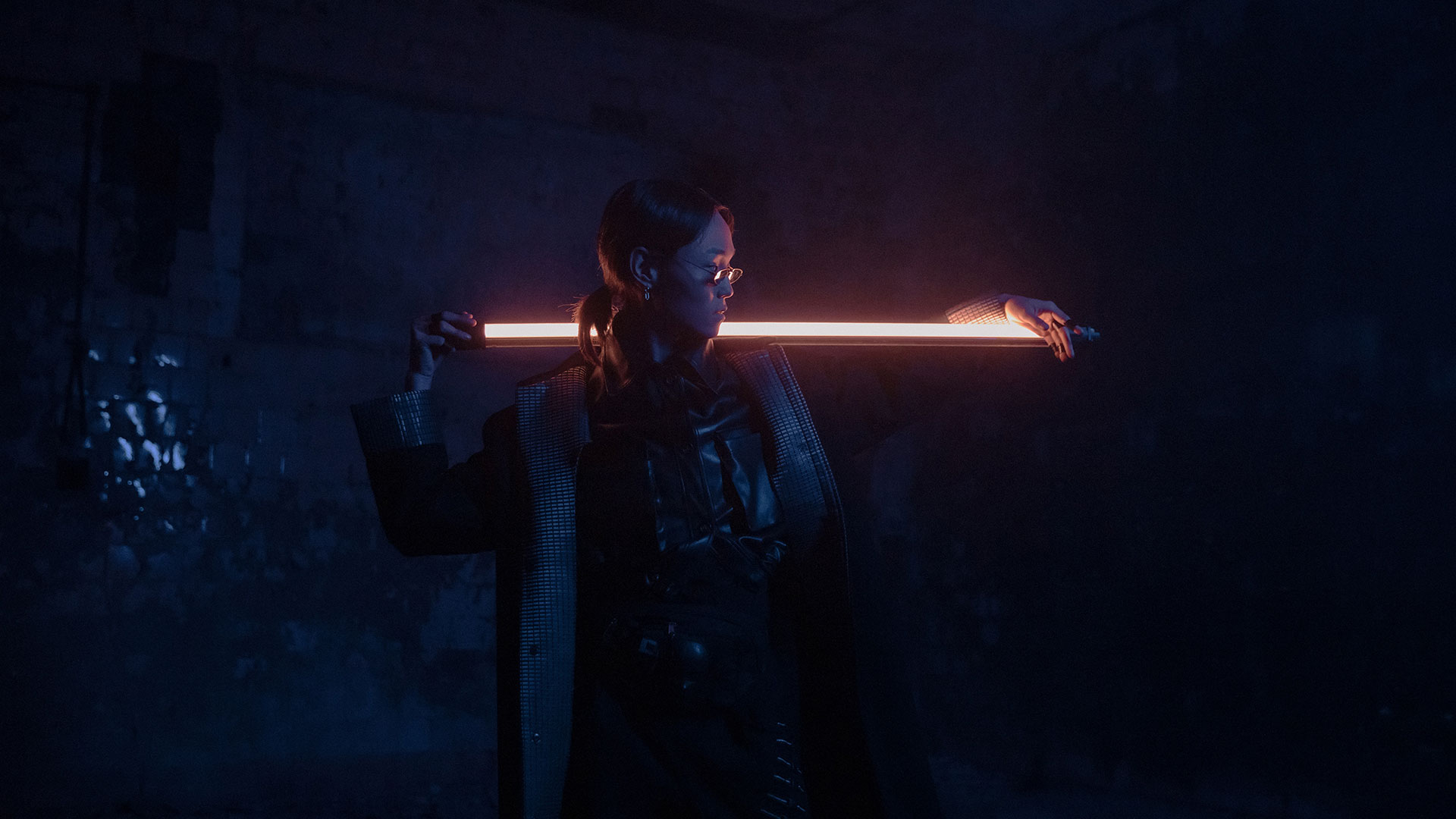 A woman wearing hip glasses and a leather jacket, holding what seems like a glowing off white sword or light saber.